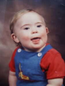 A picture of Jonathan in blue dungarees with red t-shirt on smiling aged 3.